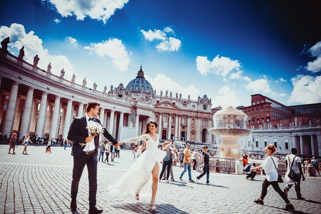 Muž v smokingu a žena v svadobných šatách sa držia za ruky a kráčajú po meste.jpg